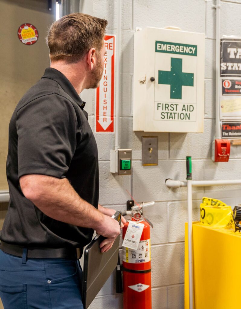A person stands in an industrial setting, looking at a fire extinguisher next to a marked First Aid Station and various safety notices on the wall.
